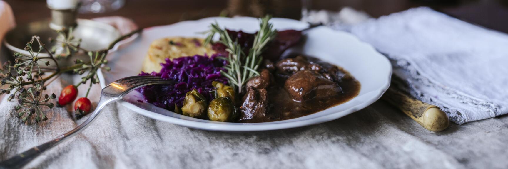 Teller mit Wildragout, Rotkohl und Knödel, Rotwein und Kerze auf gedecktem Tisch.