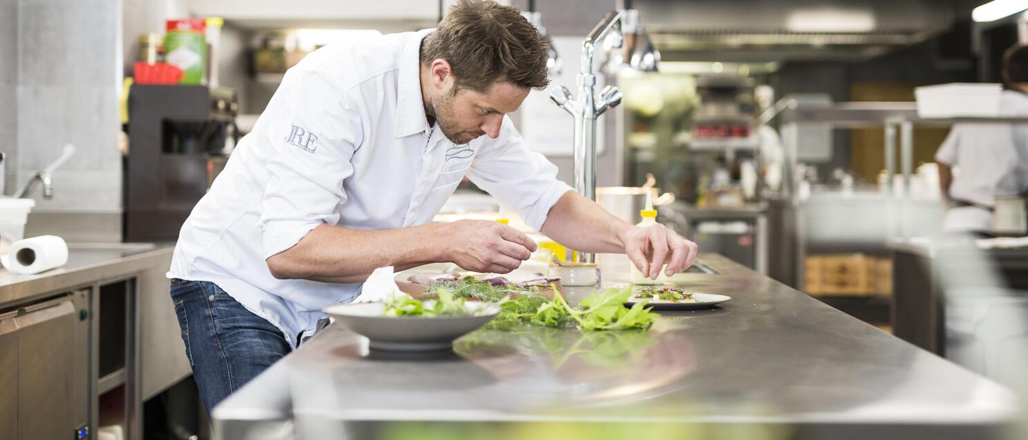 Chef in white shirt garnishing plate with fresh herbs at stainless steel workstation in professional kitchen
