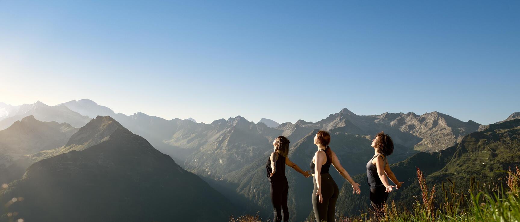 Three women in black sportswear standing on a mountain meadow with mountain panorama in the background.