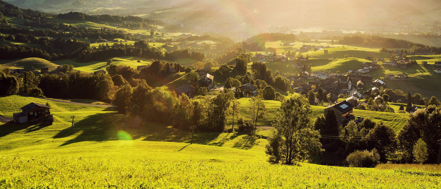 Weites Tal mit grünen Wiesen, Dorf mit Kirchturm und Bergen im Gegenlicht der Abendsonne.