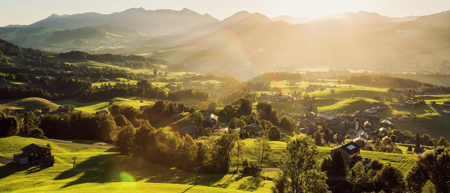 Expansive valley with green meadows, village with church spire and mountains backlit by evening sun.