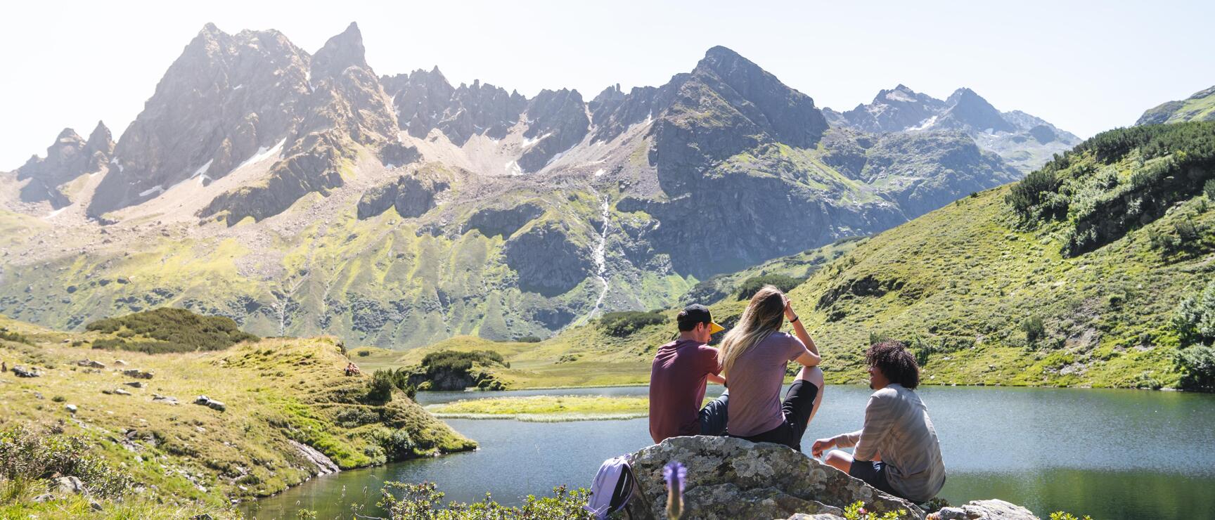 Three people sitting on a rock by an alpine lake with rocky peaks and green slopes in the background.