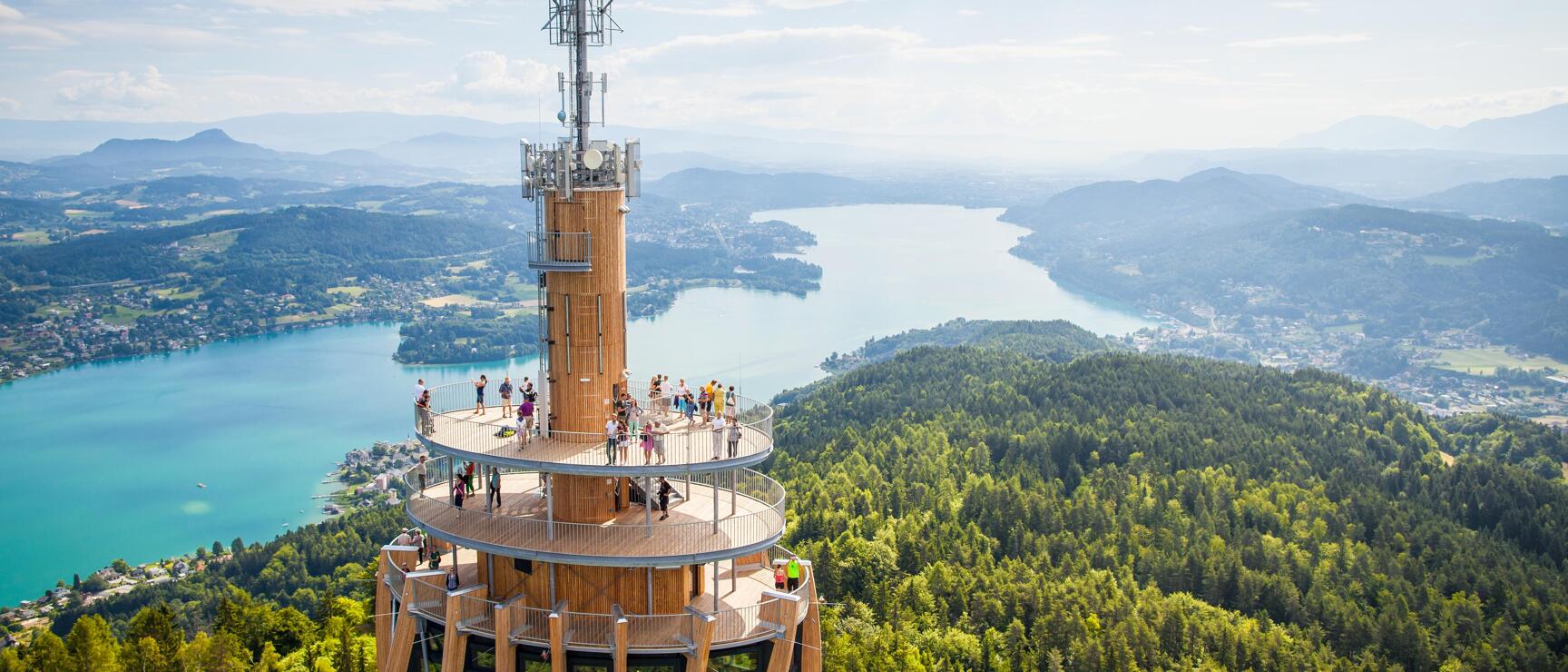 Wooden observation tower on Pyramidenkogel with visitors, Lake Wörthersee and mountain panorama in the background.