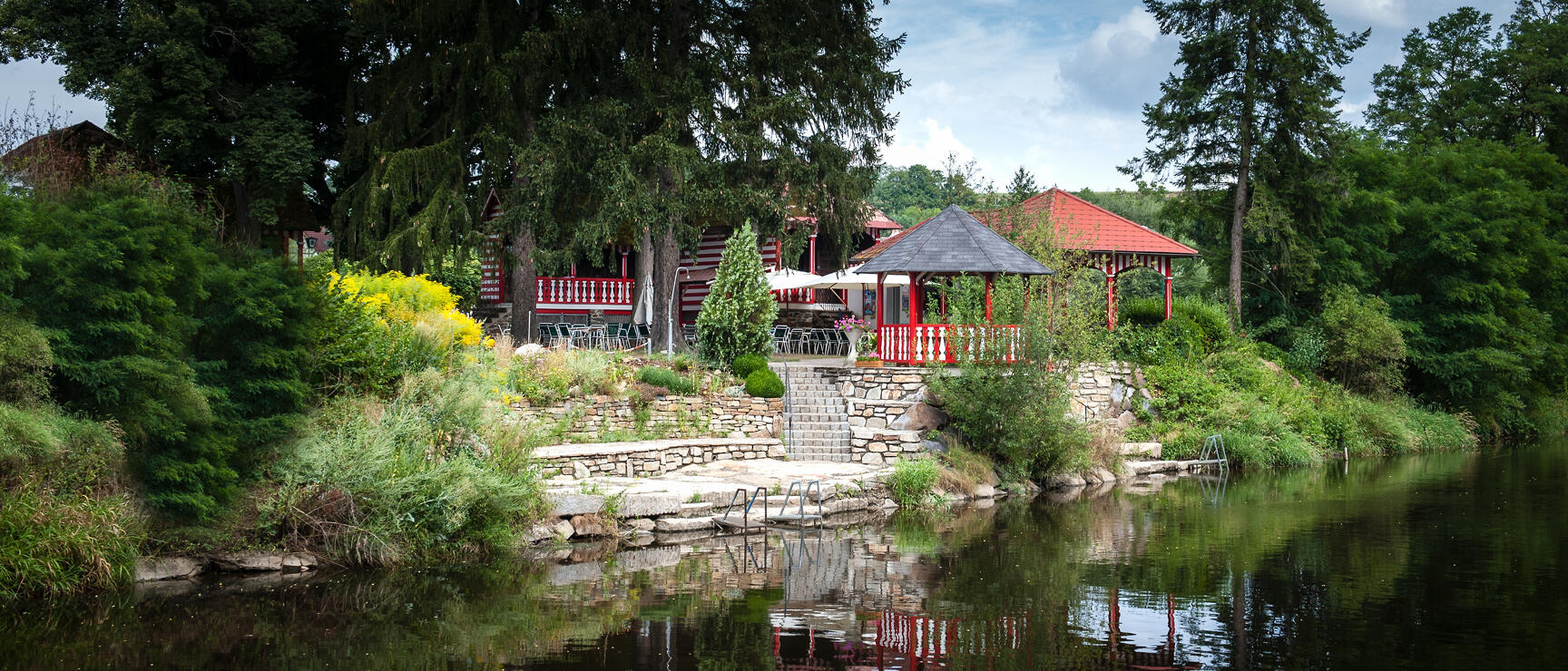 Pavilion with red roof and terrace by a pond, surrounded by trees and green plants.