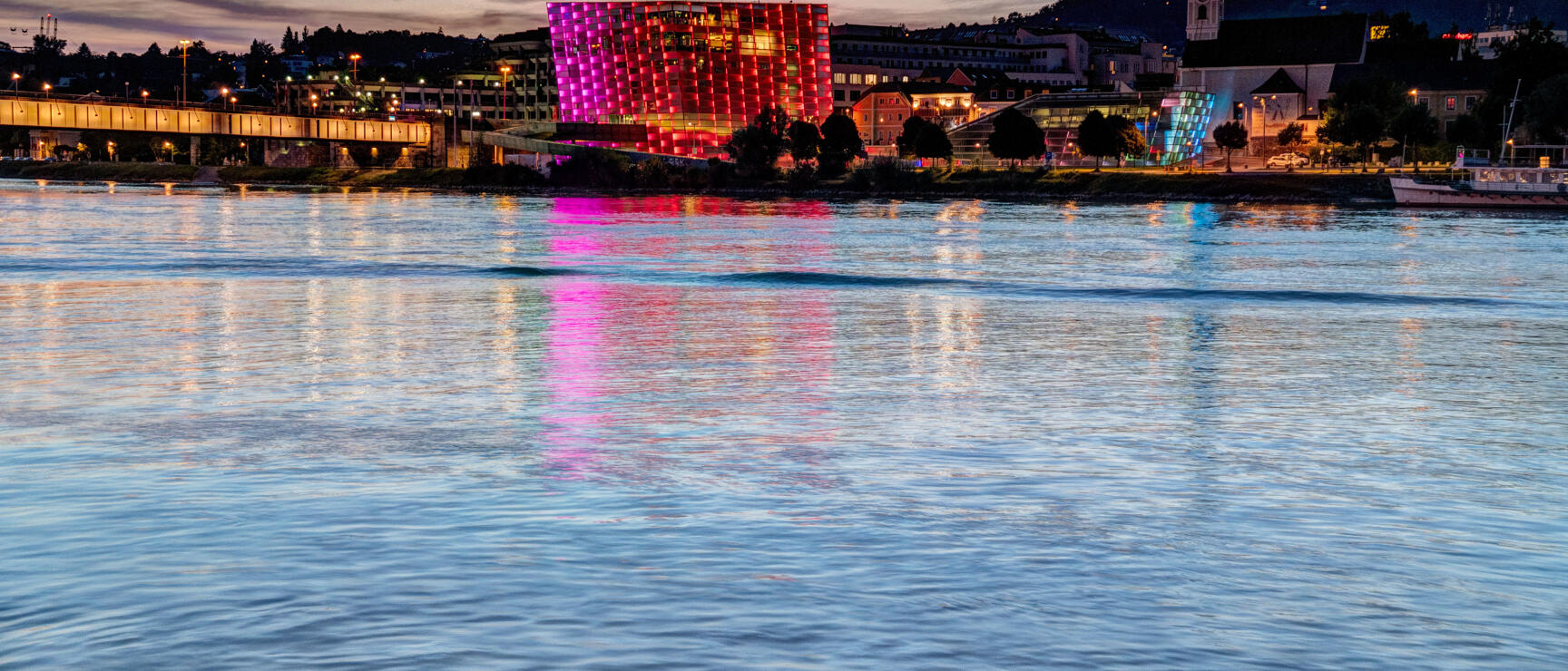 Ars Electronica Center in Linz at night, illuminated in pink-red, reflection in the Danube, church tower in the background.