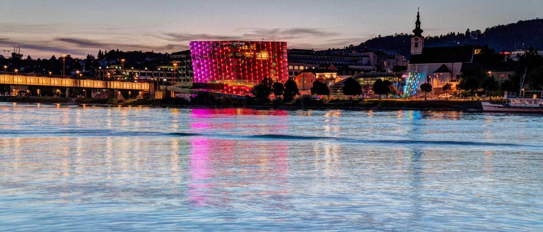 Ars Electronica Center in Linz at night, illuminated in pink-red, reflection in the Danube, church tower in the background.