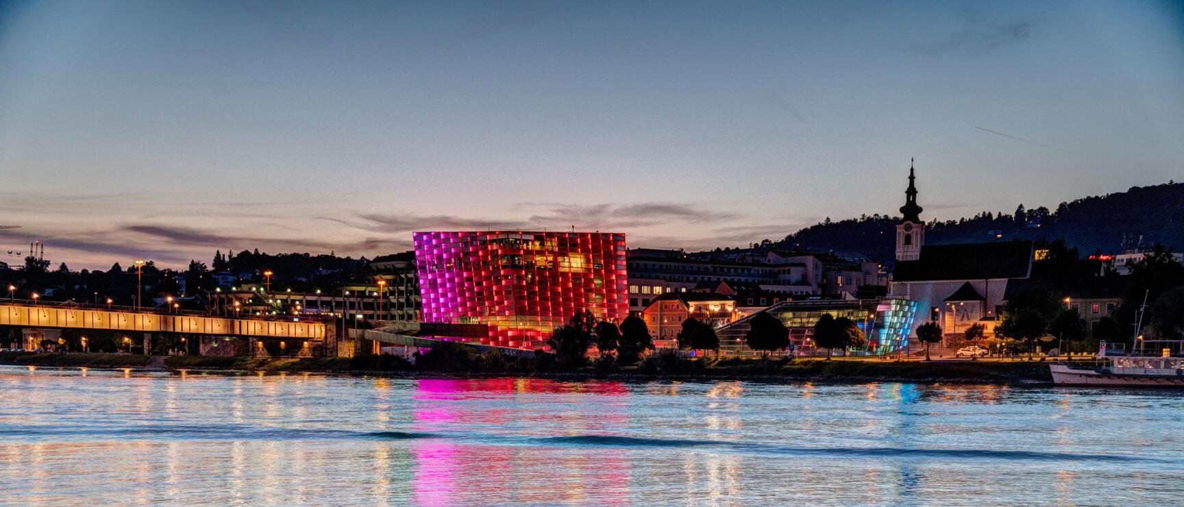 Ars Electronica Center in Linz bei Nacht, pink-rot beleuchtet, Spiegelung in der Donau, Kirchturm im Hintergrund.