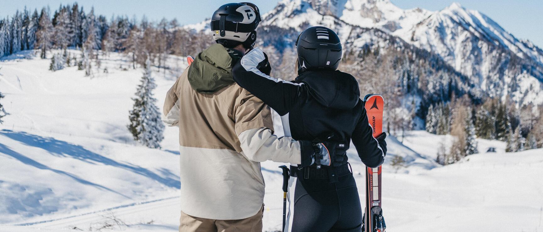 Two skiers with helmets and Atomic skis overlooking snow-covered mountain peaks and winter forest.