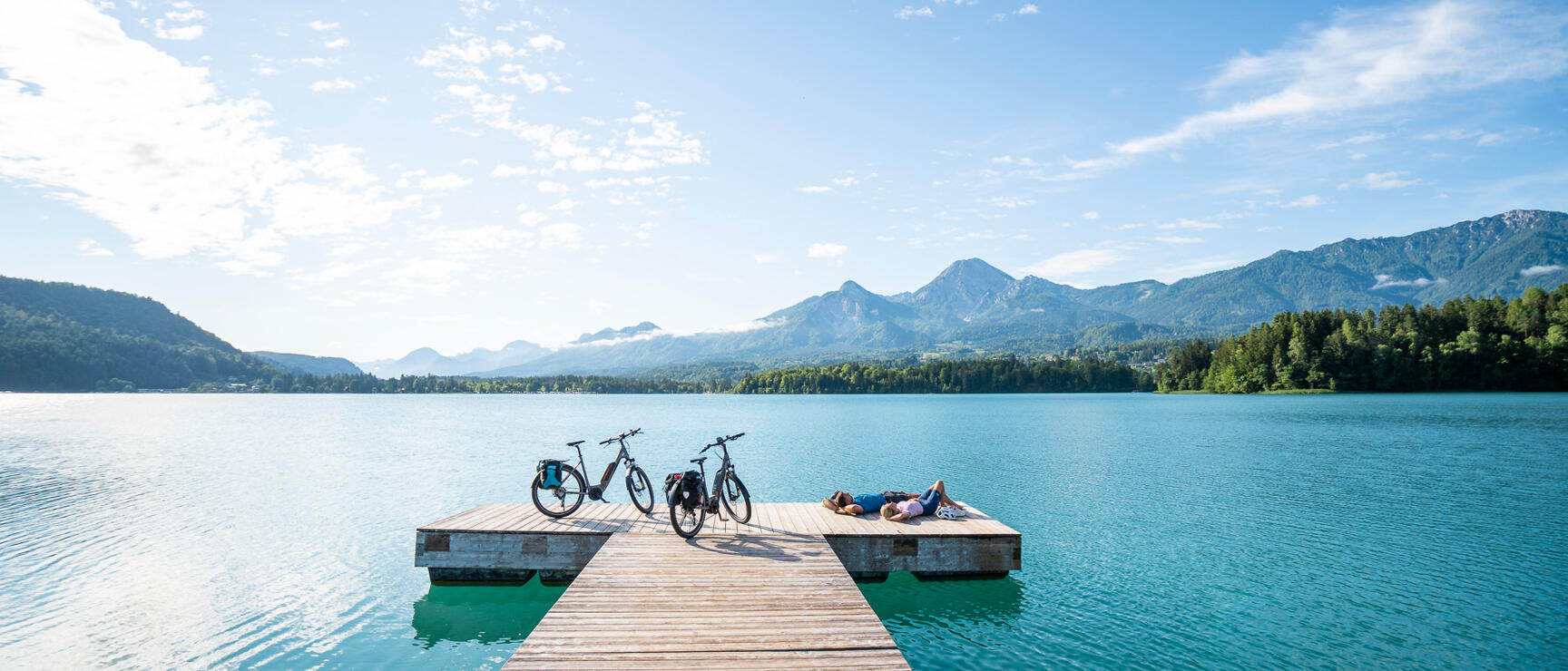 Two people lying on a wooden jetty at a turquoise lake, two bicycles beside them, mountain panorama in the background.