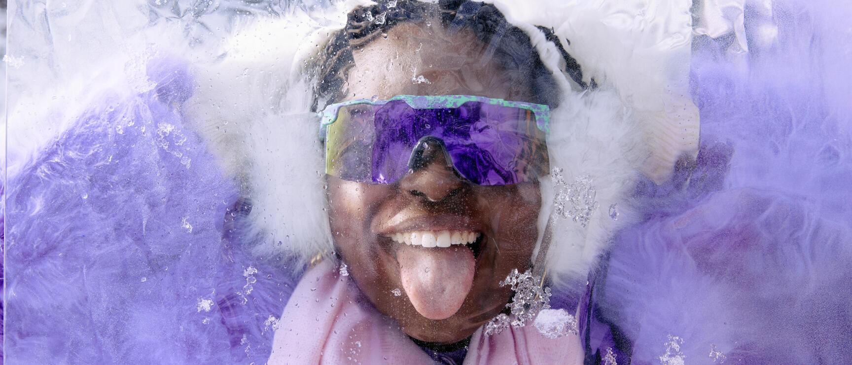 Person with purple mirrored sunglasses and ear warmers sticks tongue out, surrounded by snow and ice.