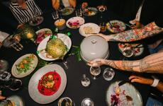 Set table with colourful dishes, wine glasses and tattooed arms serving food.