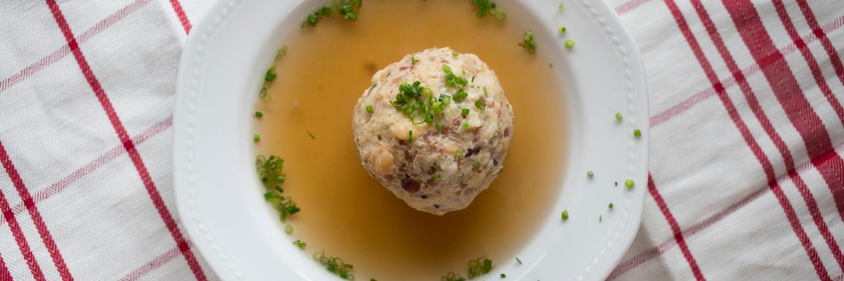 Liver dumpling soup in white bowl with chives, on red and white kitchen towel, overhead view.