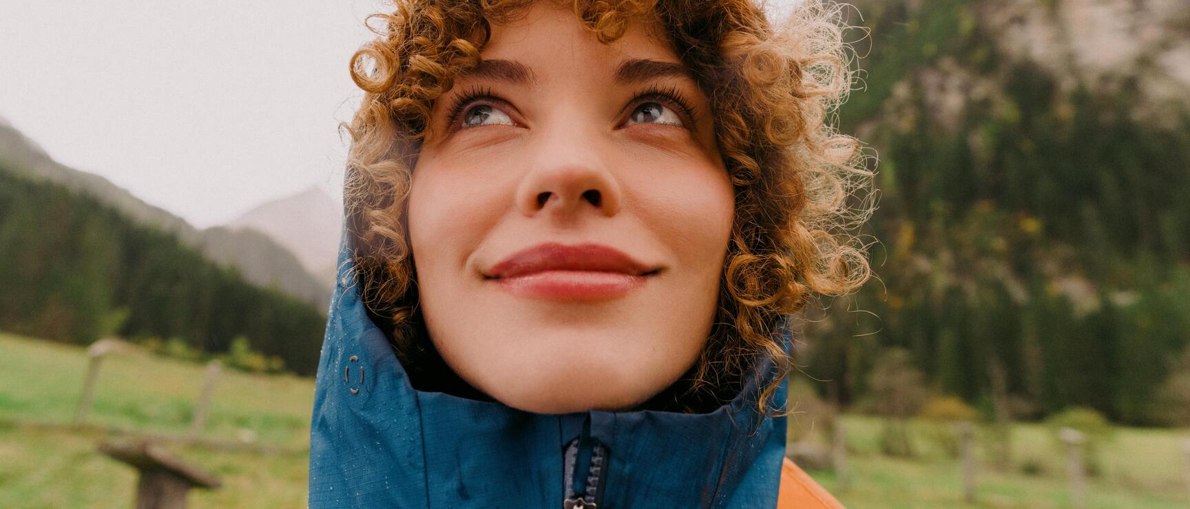 Woman with curly hair in blue rain jacket looking upward, green mountain meadow in background.