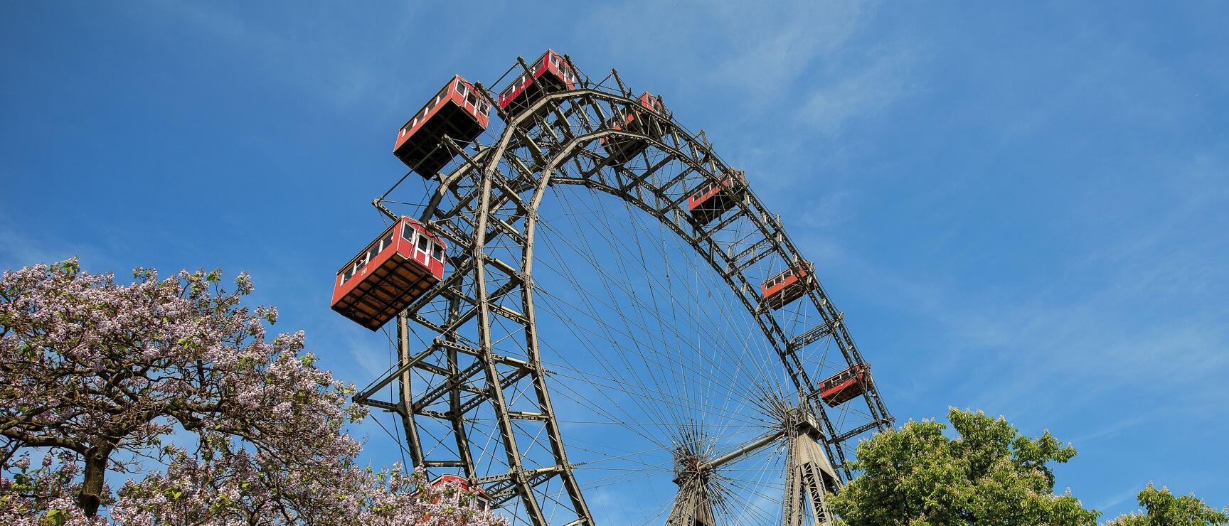 Vienna's Ferris wheel in the Prater with red gondolas, blooming trees in foreground, blue sky.