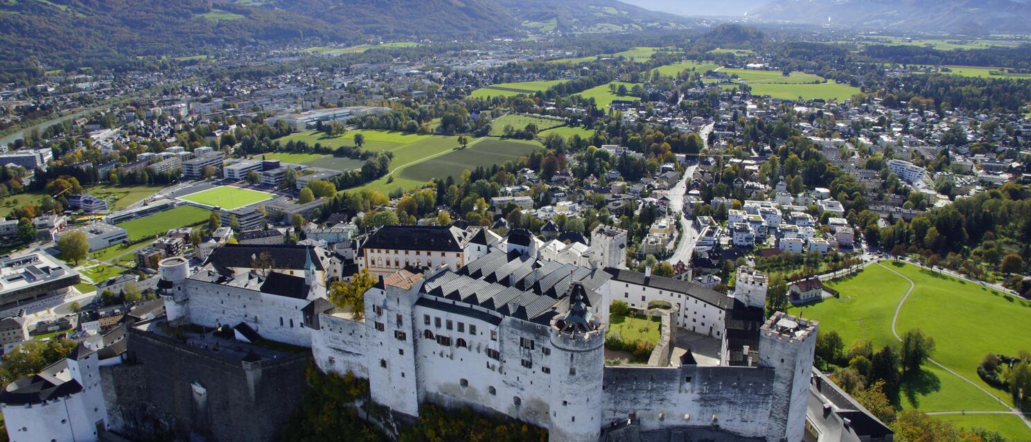 Aerial view of Hohensalzburg Fortress with city of Salzburg and alpine panorama in the background.