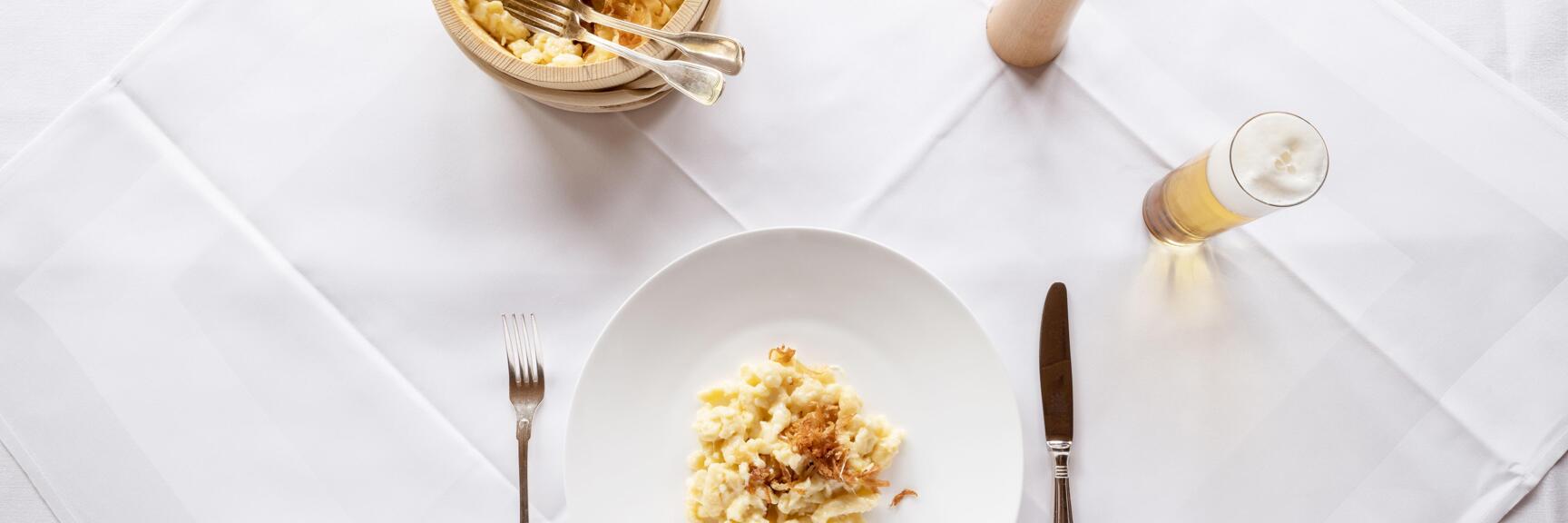 Cheese spätzle with crispy onions on white plate, wooden bowl, pepper mill and beer glass from above.
