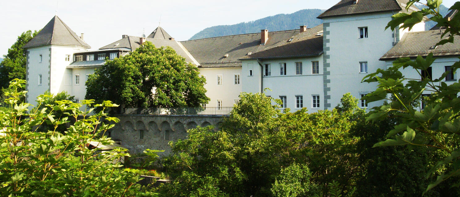 White castle with corner towers, surrounded by trees, stone wall in foreground, mountain in background.