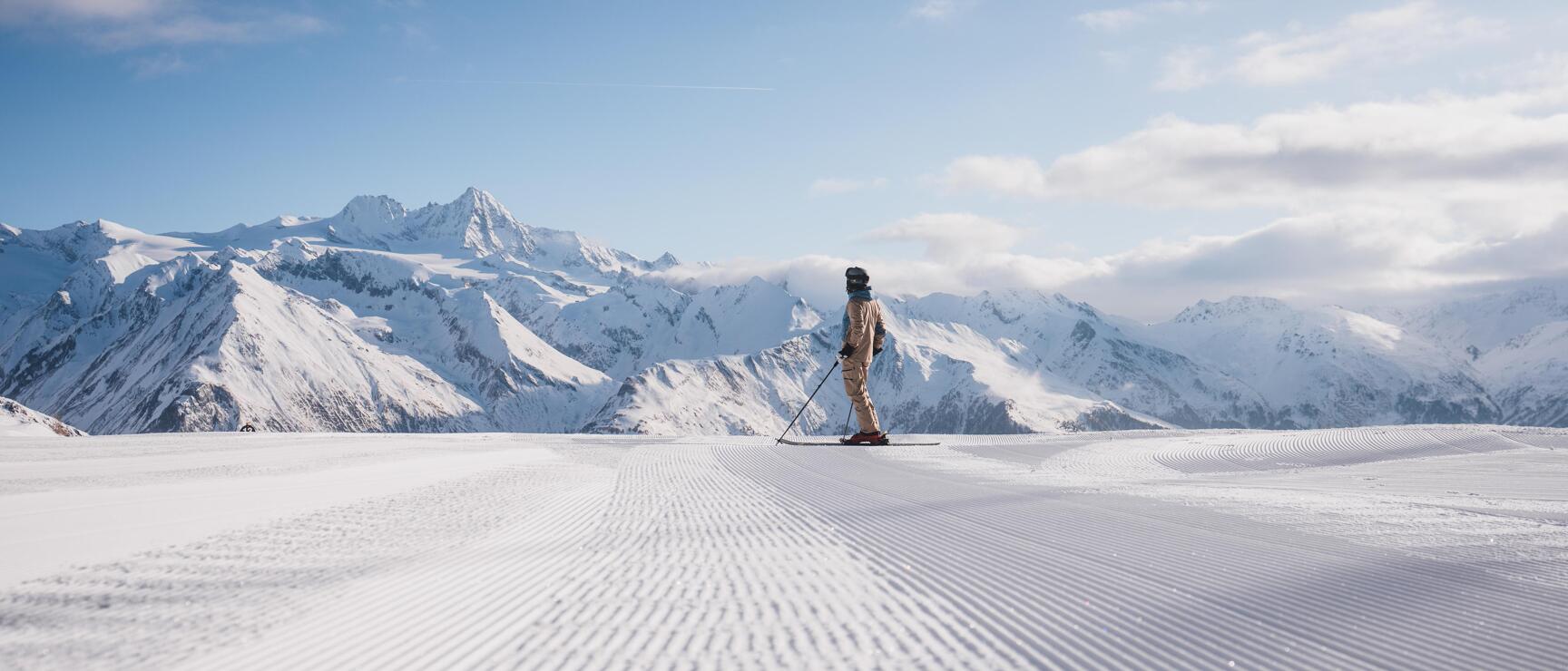 Skier on a prepared slope with a view of snow-covered Alpine peaks under a blue sky.