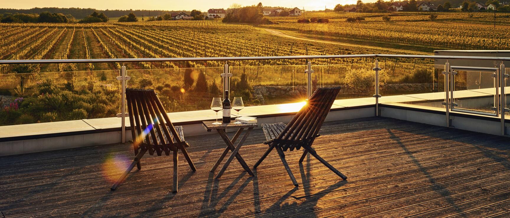 Terrasse mit zwei Holzstühlen, Weinflasche und Gläsern, Blick auf Weinberge im Abendlicht.