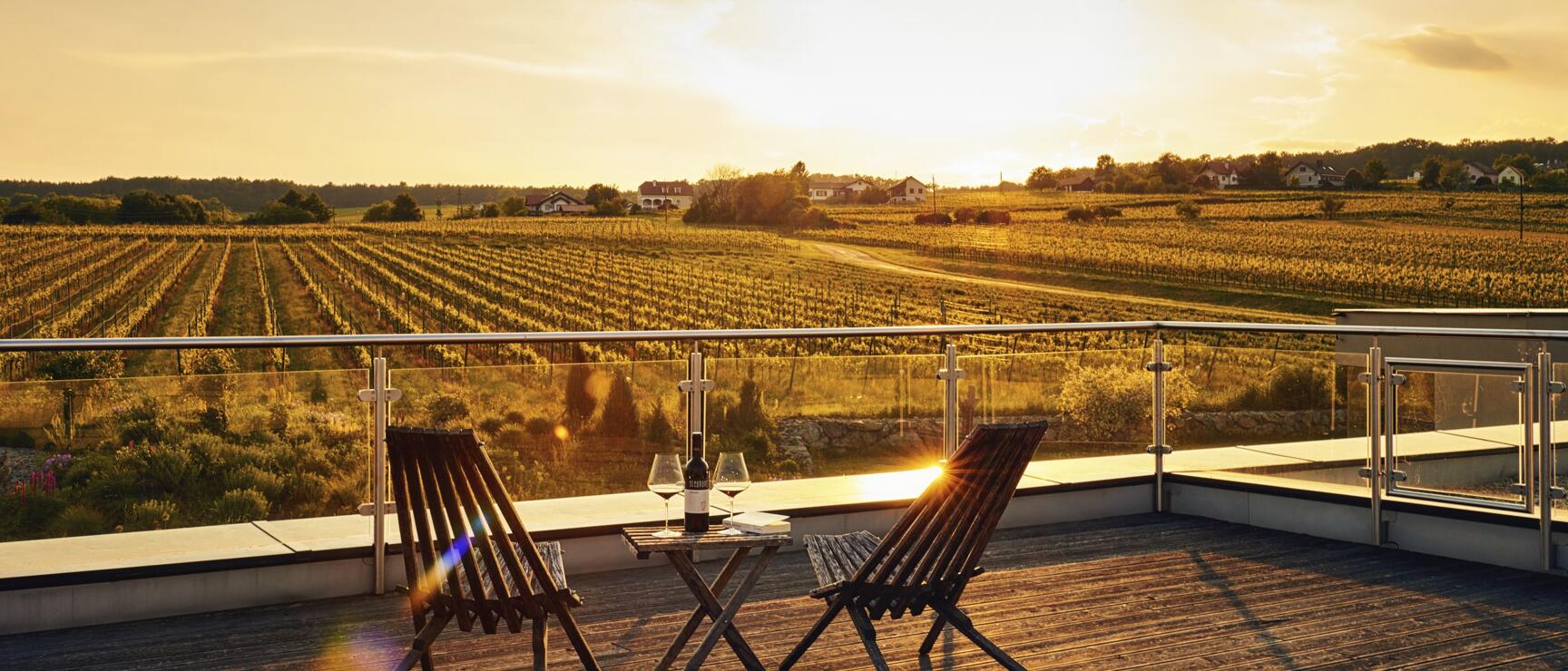 Terrace with two wooden chairs, wine bottle and glasses, view of vineyards in evening light.