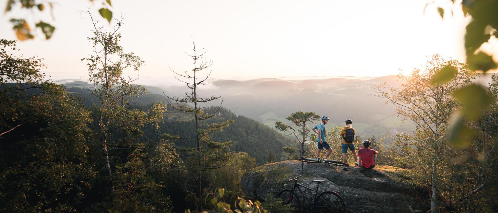 Three mountain bikers with helmets resting on a rock, bikes beside them, distant forest view in evening light.