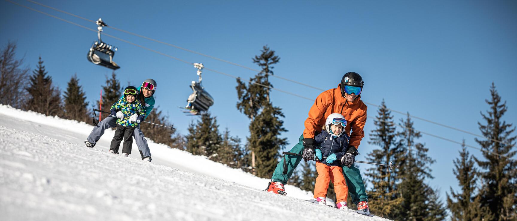 Two adults each with a toddler skiing on a sunny slope with chair lift in the background.