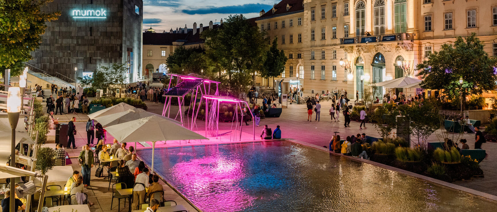 MuseumsQuartier Vienna at night, pink light installation at the water basin, mumok and illuminated Baroque building in the background.