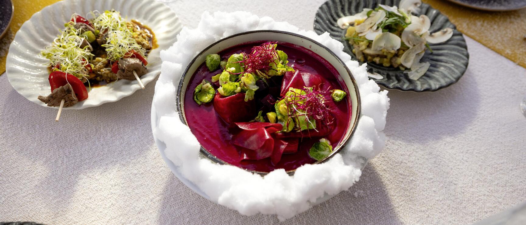 Beetroot soup with sprouts in a bowl on cotton, flanked by two appetizers on a set table.