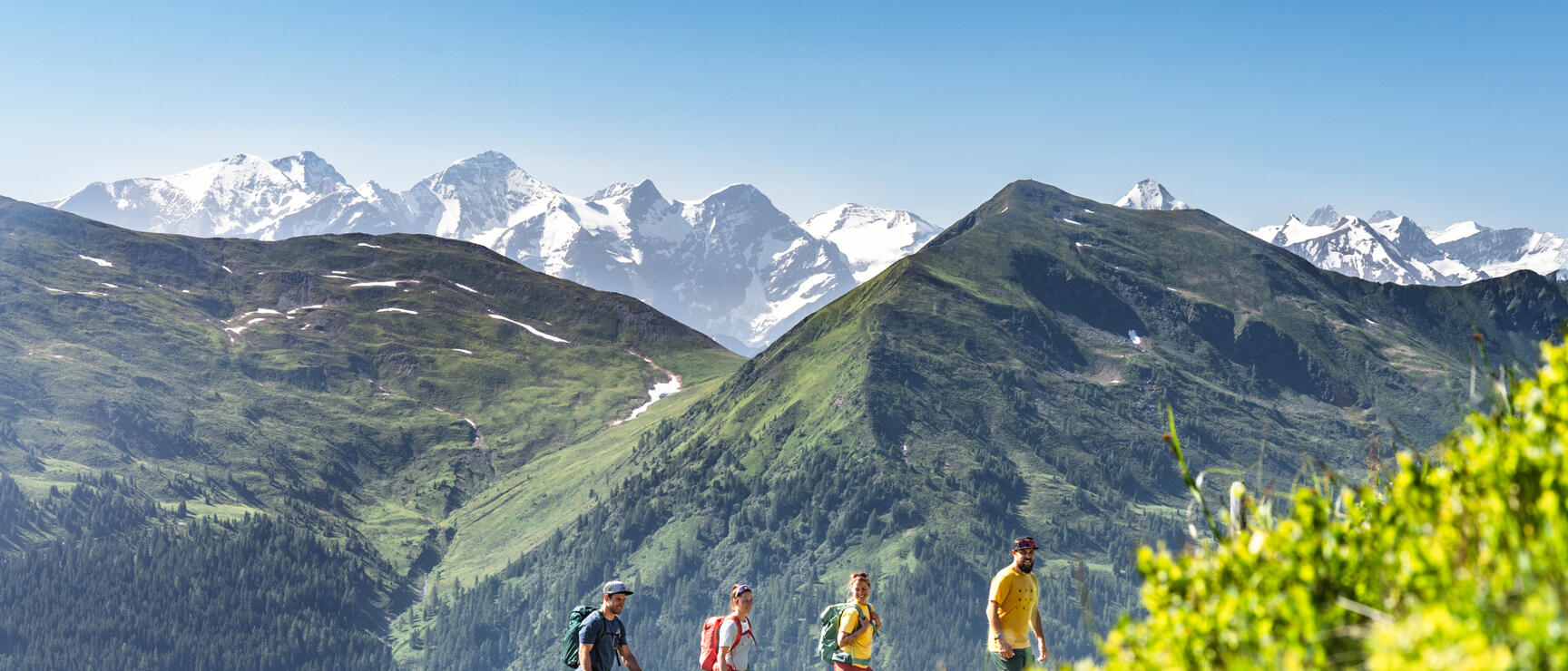 Four hikers with backpacks on a mountain trail, snow-covered Alpine peaks in the background.