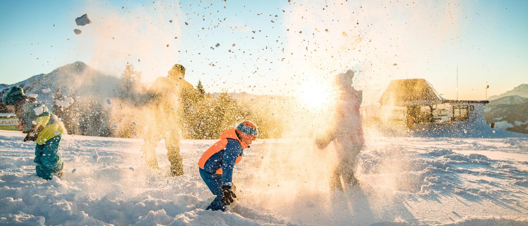 Children and adults having a snowball fight backlit by the evening sun, mountains and cabin in the background.