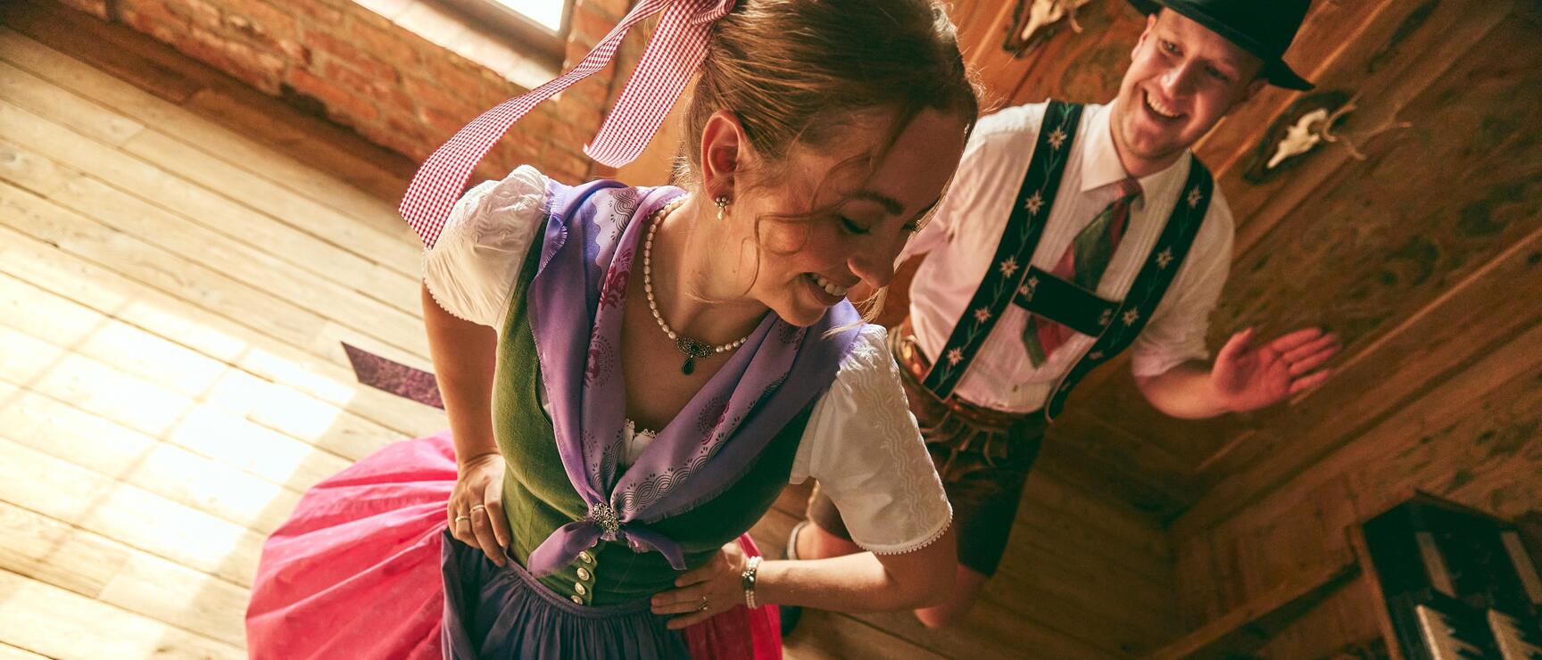 Woman in dirndl and man in traditional costume laughing while dancing on wooden floor in rustic room.