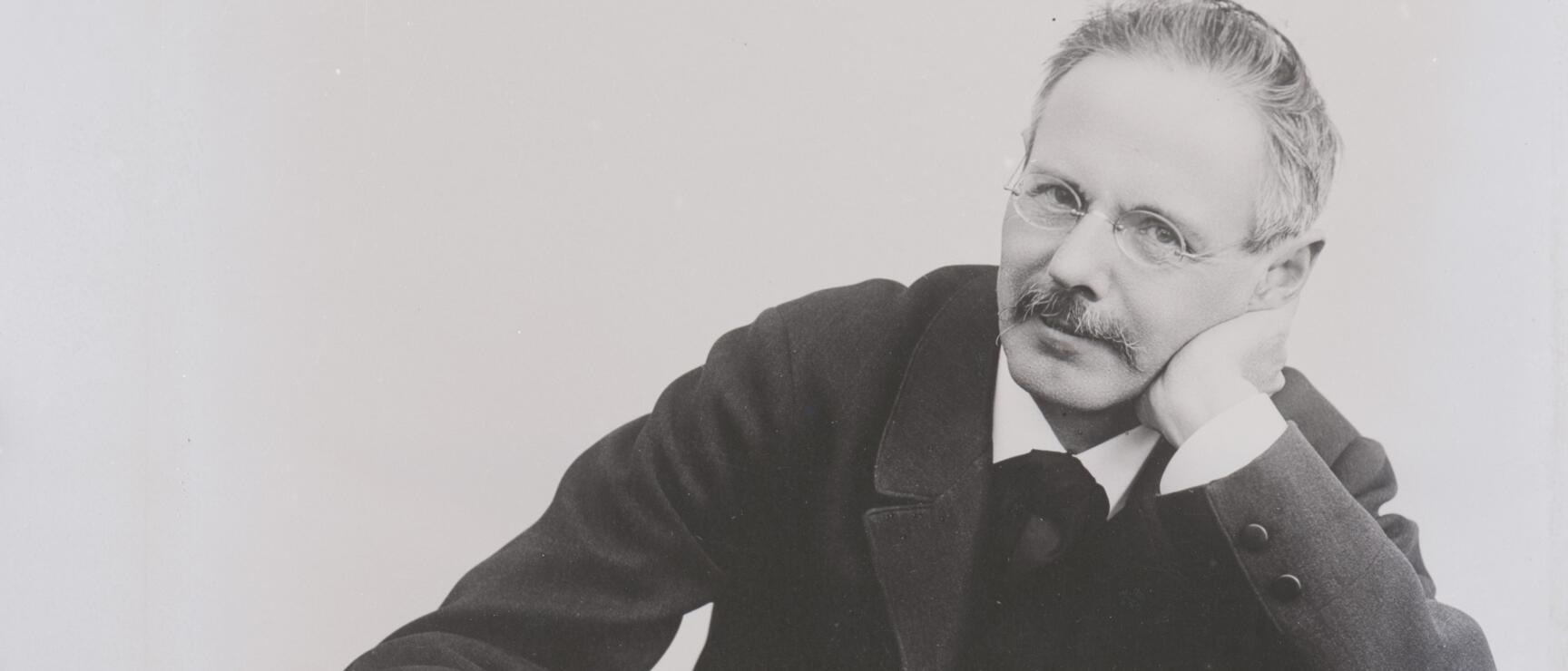 Black and white portrait of a man with glasses and moustache, sitting at a table with a book, head resting on hand.