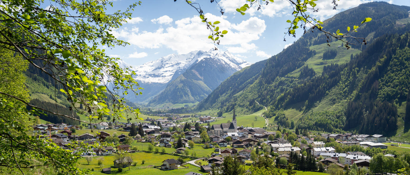 Alpine village with church in the valley, green meadows, forests and snow-capped peaks in the background.
