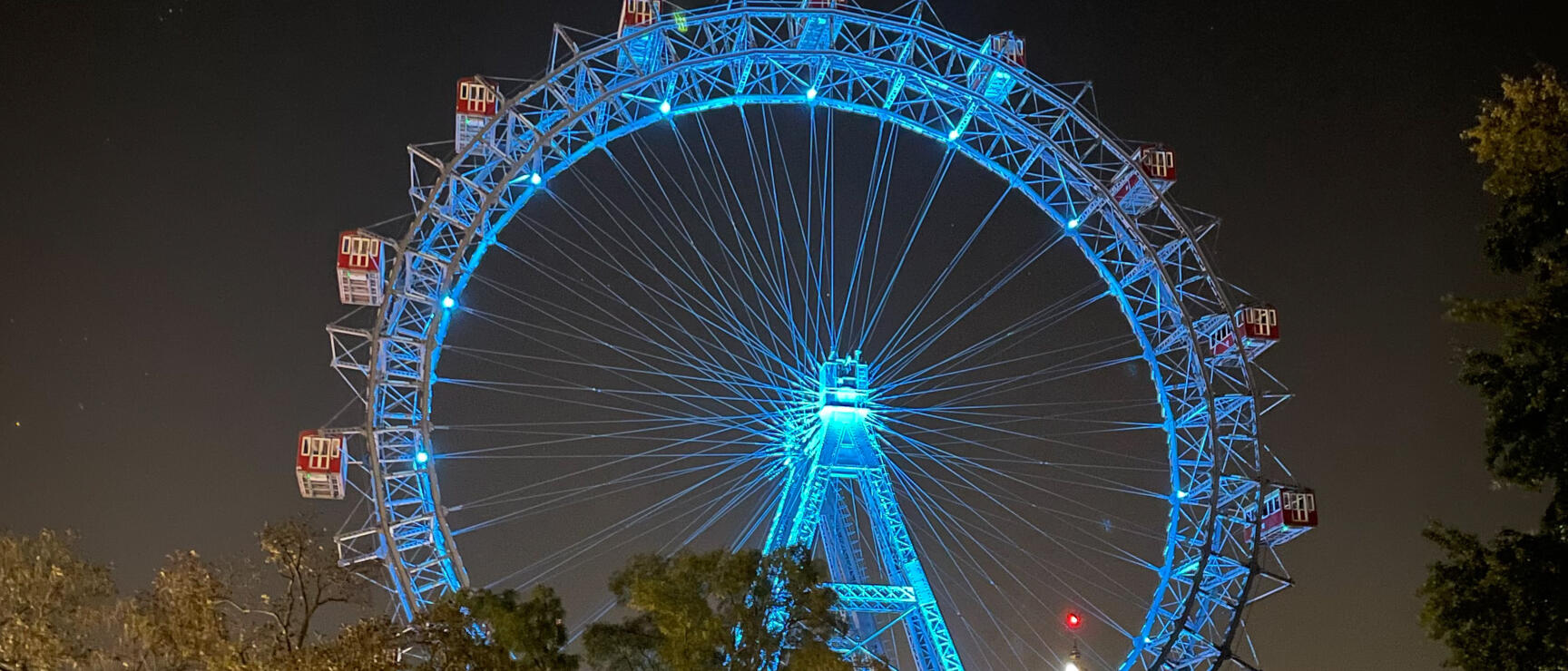 Vienna Giant Ferris Wheel at night, blue illuminated, red gondolas, park with trees and street lamp in foreground.