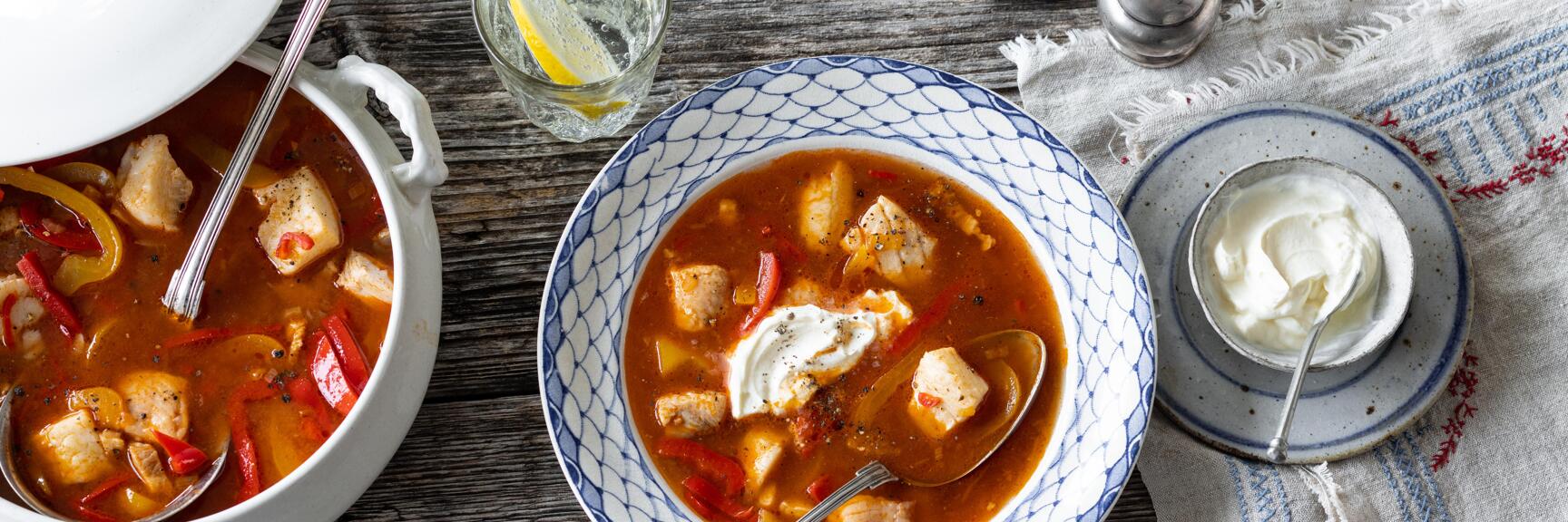 Fish goulash with paprika in blue and white bowl, sour cream, glass of water with lemon on wooden table.