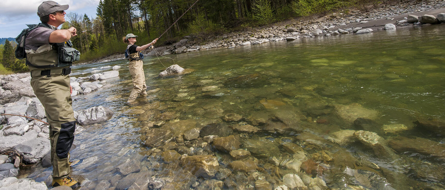 Two people in wading gear fly fishing in a clear mountain river, coniferous forest in the background.
