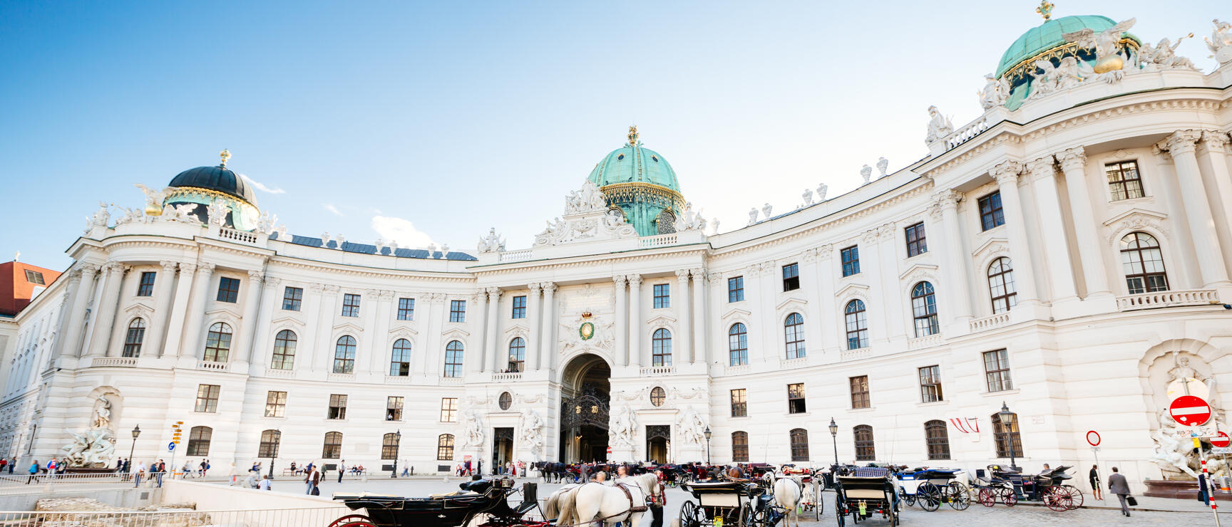 Fiaker mit weißen Pferden vor der Wiener Hofburg, barocke Fassade mit grünen Kuppeln, blauer Himmel.