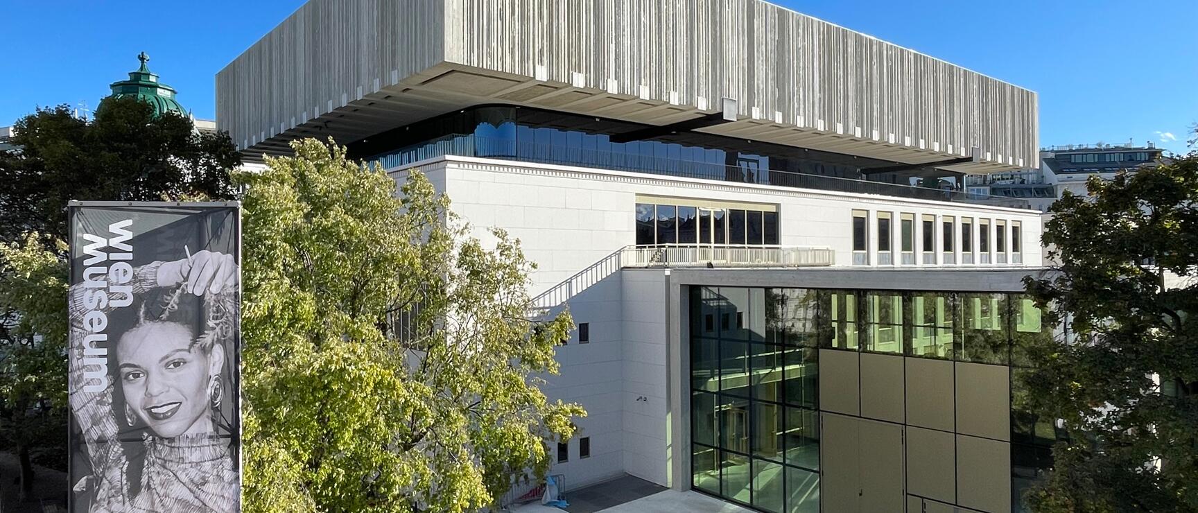 Exterior view of Vienna Museum at Karlsplatz with modern roof structure, banner with female portrait on left.