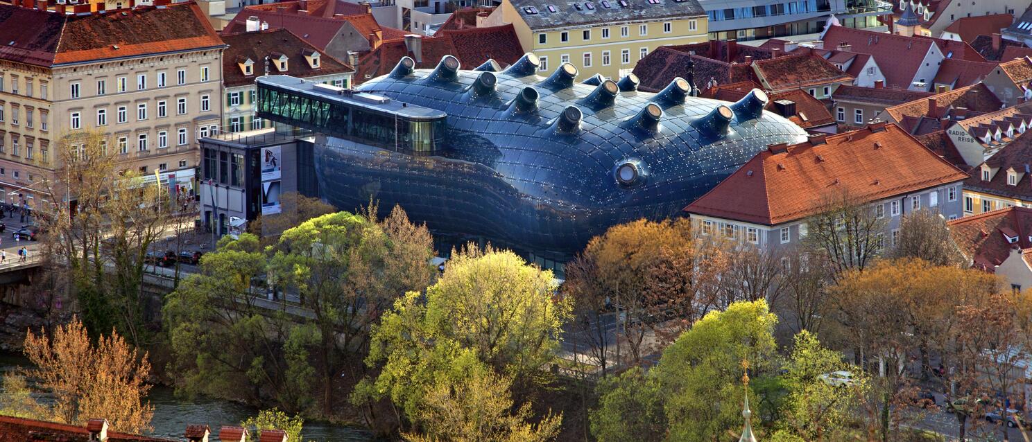 Aerial view of Kunsthaus Graz with blue biomorphic façade amid old town, Mur river, red tile roofs.