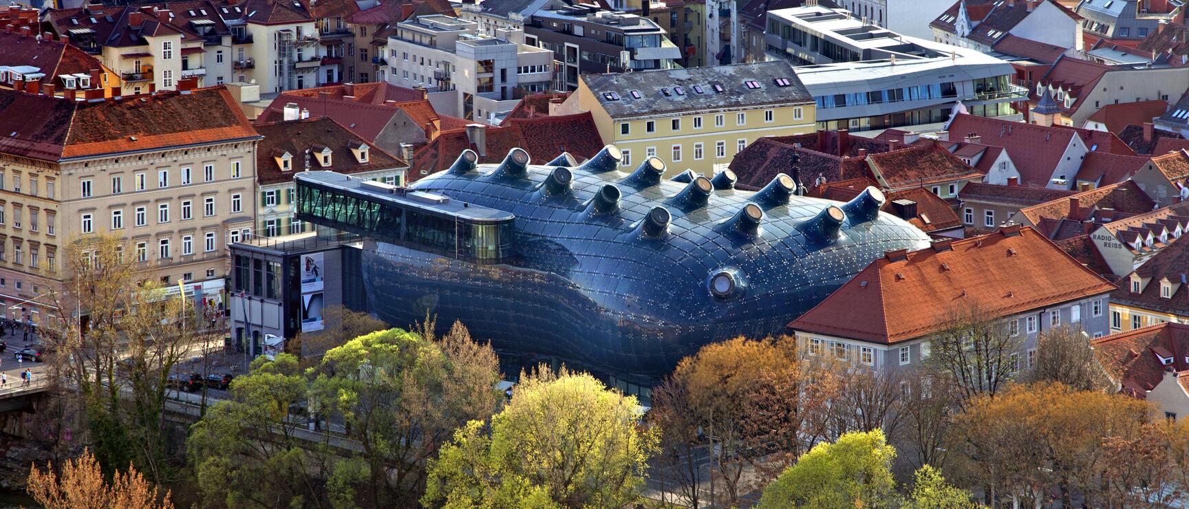 Aerial view of Kunsthaus Graz with blue biomorphic façade amid old town, Mur river, red tile roofs.