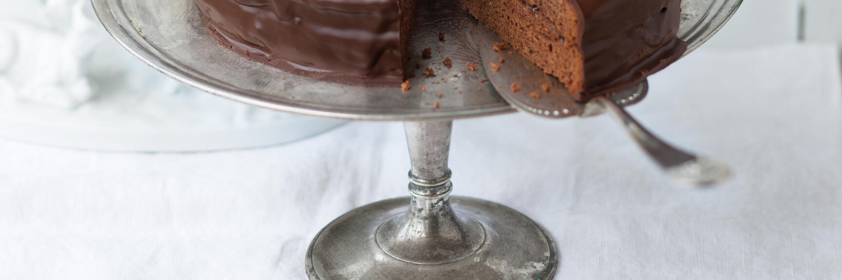 Sachertorte with chocolate glaze on silver cake plate, one slice cut, white background.