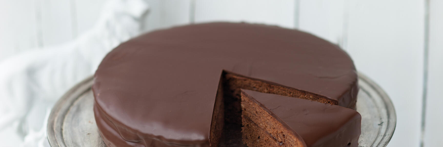 Sachertorte with chocolate glaze on silver cake plate, one slice cut, white background.