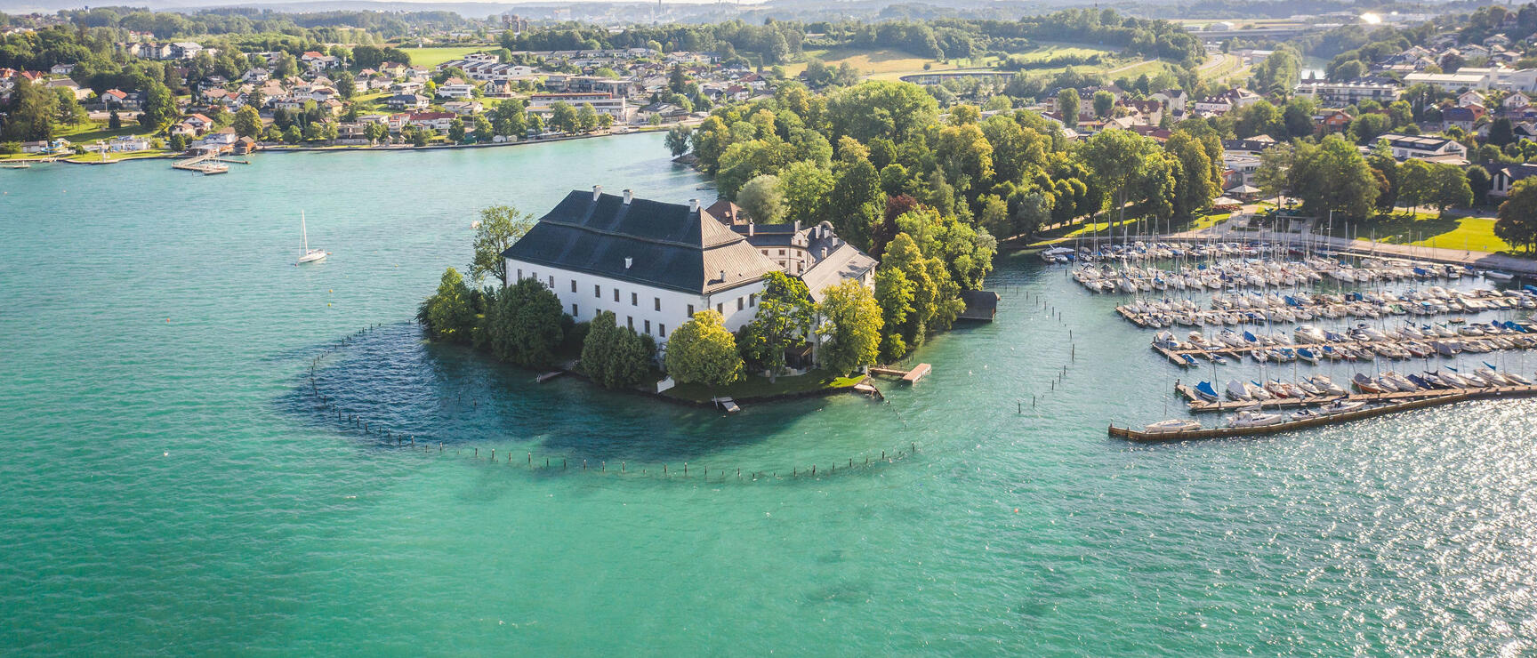 White castle on peninsula in turquoise Traunsee lake, yacht harbor on right, village in background.