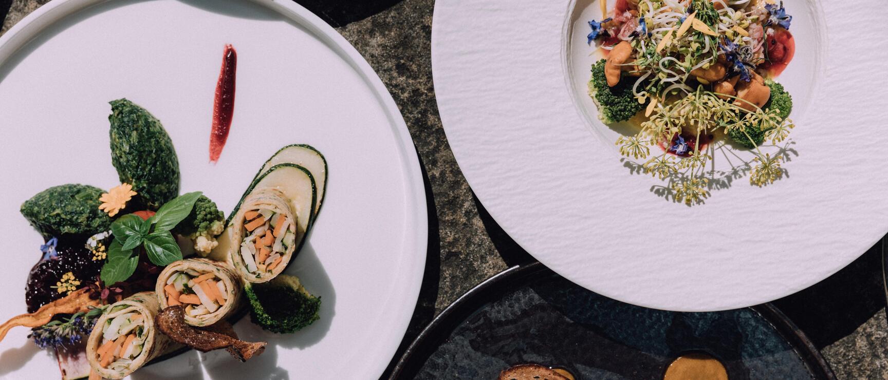 Drei Gourmet-Teller mit kunstvoll angerichteten Gemüsegerichten, Blüten und Kräutern, Draufsicht.