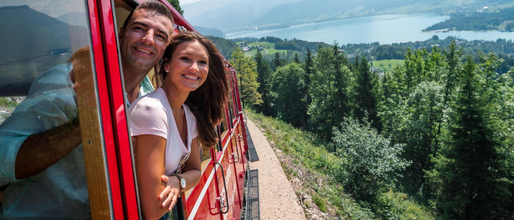 Two people leaning out of red cogwheel train with lake view and mountain panorama in background