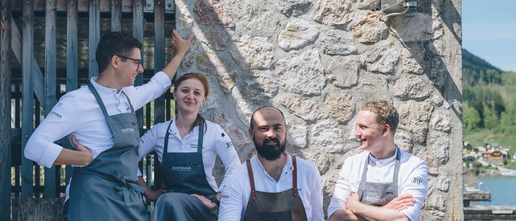 Four chefs in white chef coats and grey aprons in front of a stone wall by the lake.