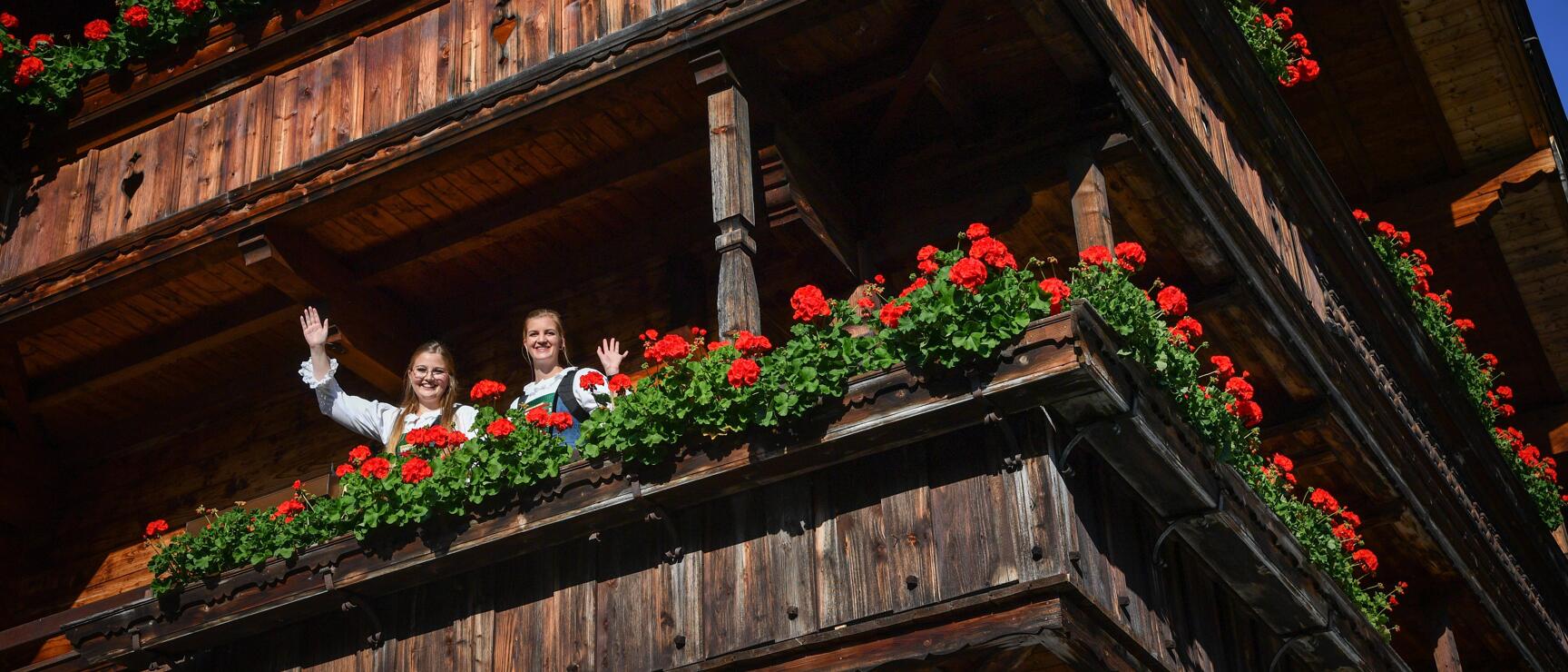 Zwei Frauen in Tracht winken von einem Holzbalkon mit roten Geranien, Schild „Zimmer frei" sichtbar.