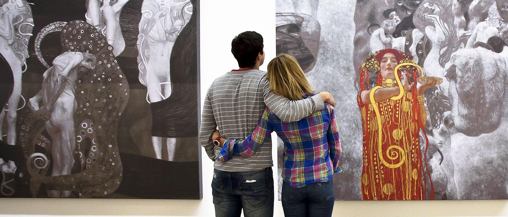 Two people viewing large-format Klimt paintings in a museum, wooden floor, white walls.