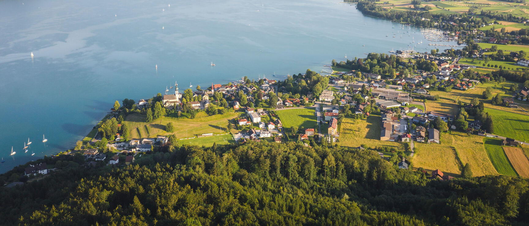 Aerial view of Lake Atter with shoreline settlement, sailing boats, and mountain range in the background.