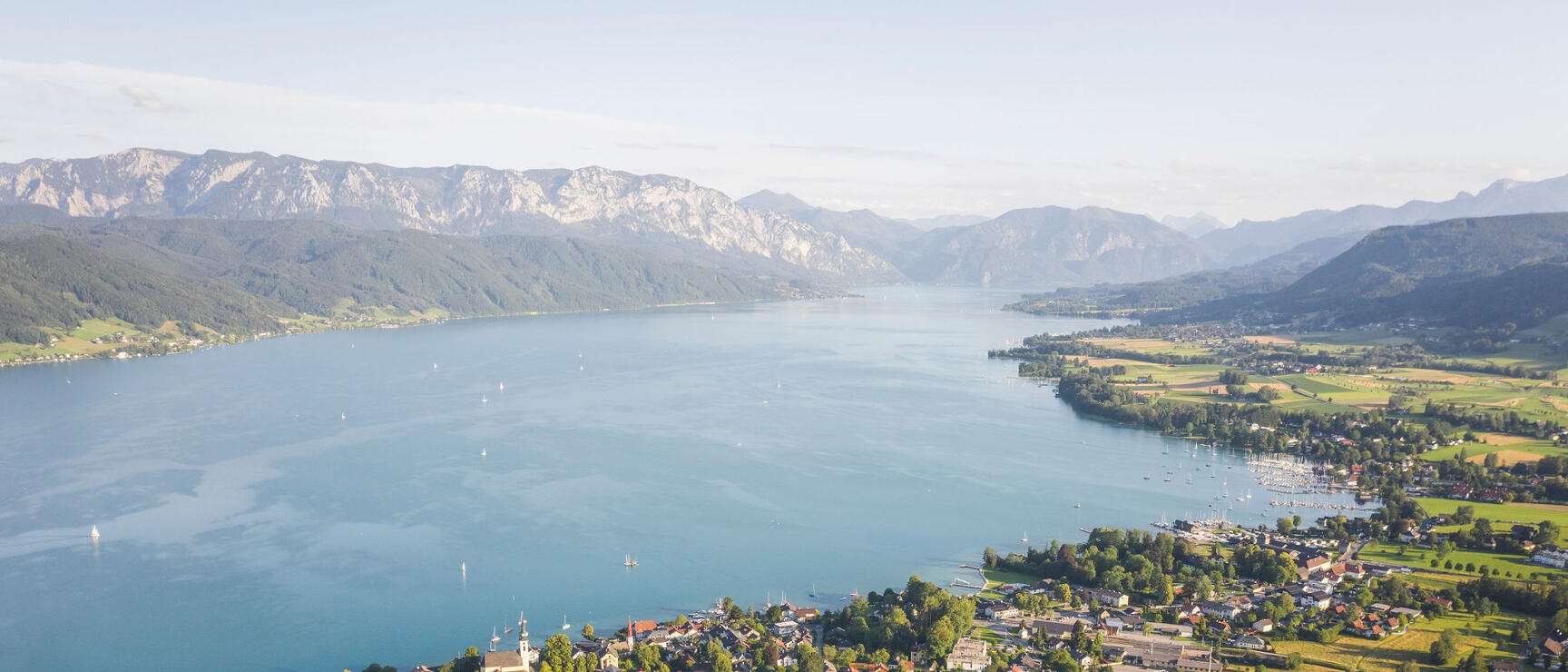 Aerial view of Lake Atter with shoreline settlement, sailing boats, and mountain range in the background.
